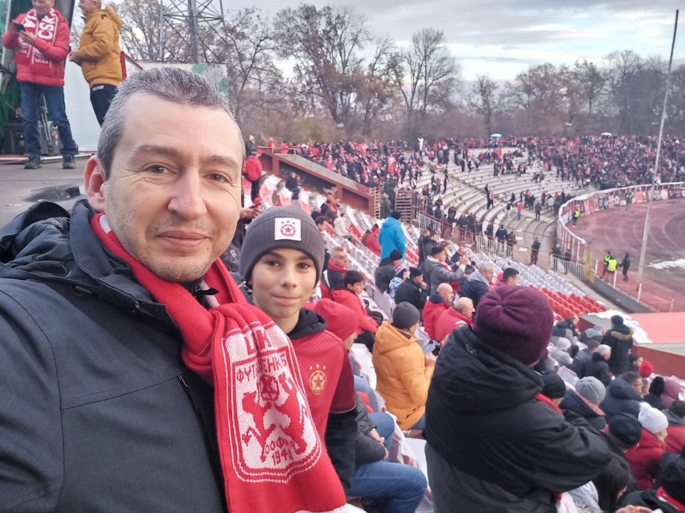 Мъж и момче в сектор А при Лудогорец; зад тях пълните трибуни и червената писта.