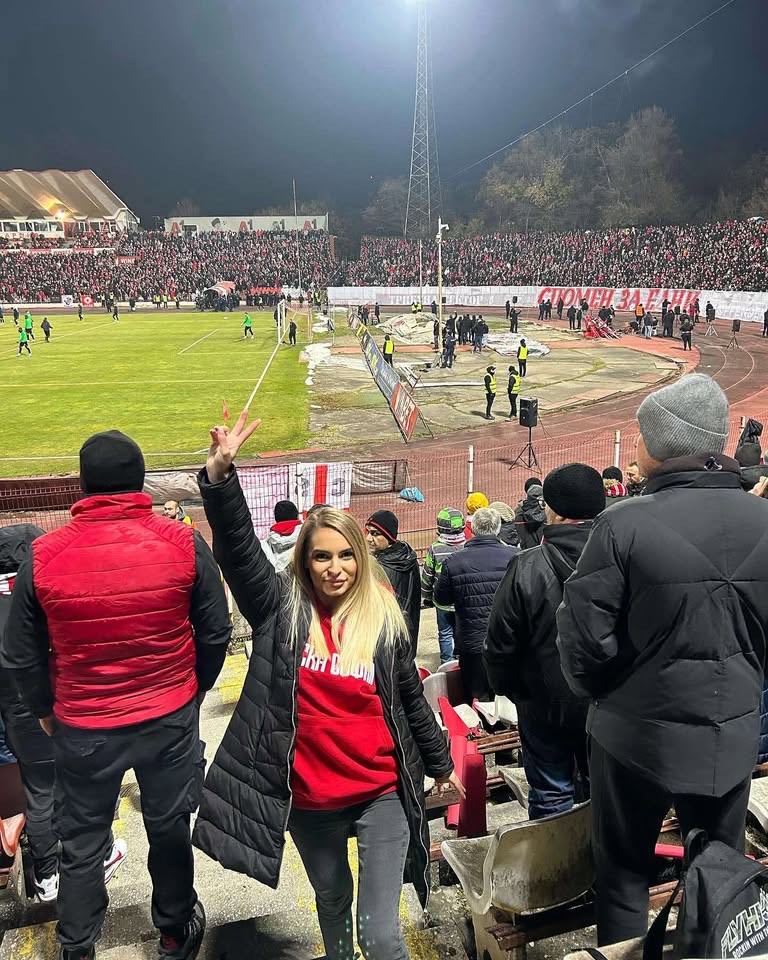 Нощ на старата Армия при Лудогорец: привърженичка в червен суитшърт по стъпалата на трибуната, зад нея пълните сектори, теренът вляво и транспаранти по оградата.